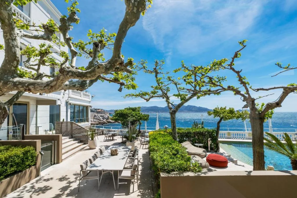 area externa do Le Petit Nice - Passedat, com uma mesa e oito cadeiras a esquerda, uma piscina a direita, arvores e um céu azul com poucas nuvens e o mar de Marselha ao fundo