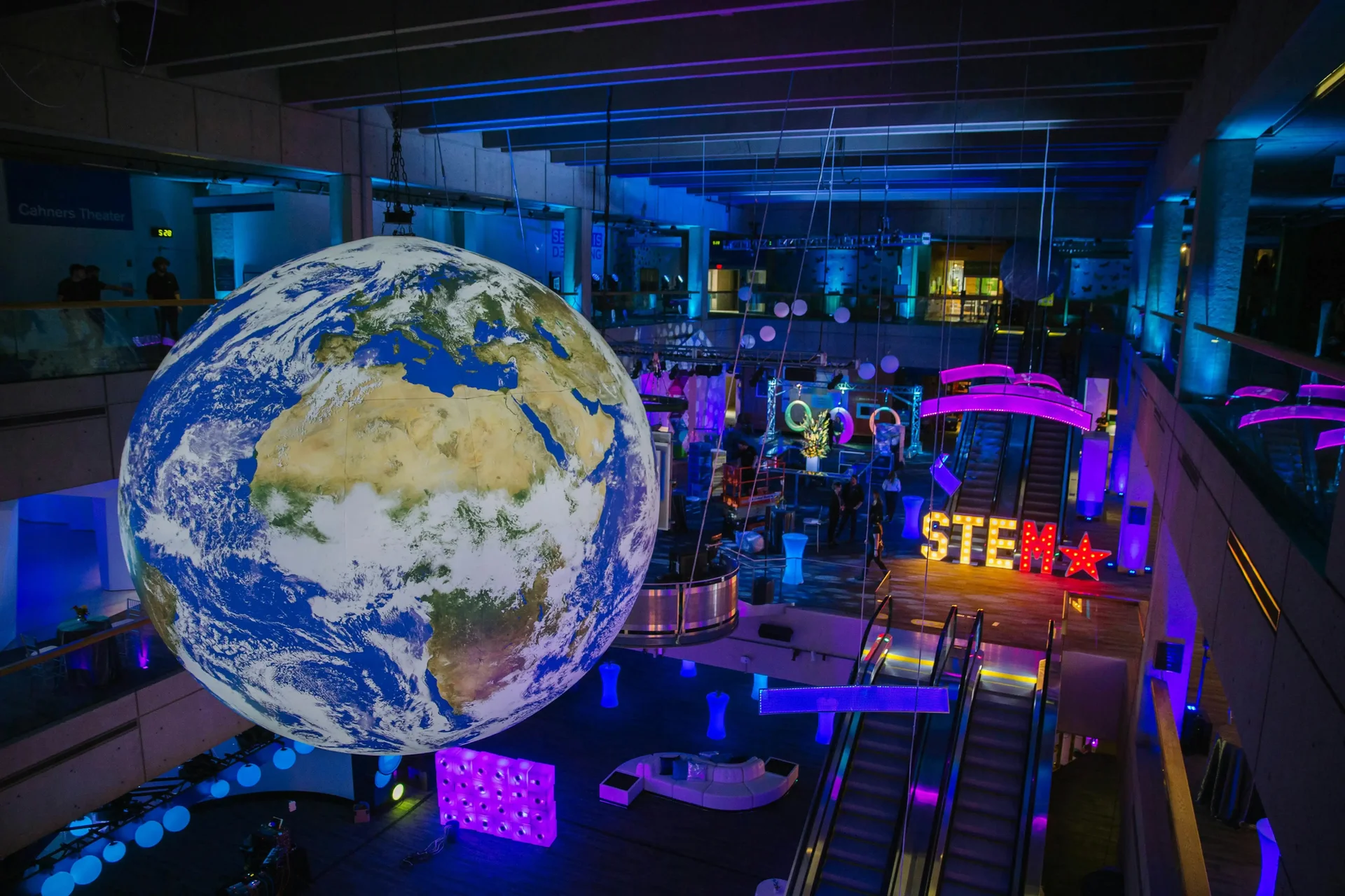 Exposição no Museu da Ciência de Boston com uma grande réplica do planeta Terra suspensa, rodeada por luzes coloridas e decorações inspiradas na temática STEM (Ciência, Tecnologia, Engenharia e Matemática).