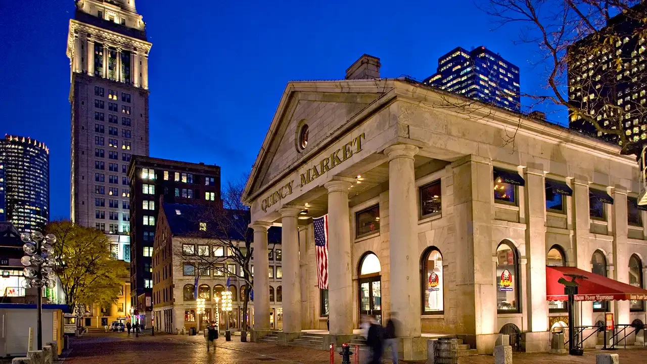 Quincy Market em Boston à noite, com iluminação encantadora e uma bandeira dos EUA na entrada, criando uma cena charmosa e histórica.