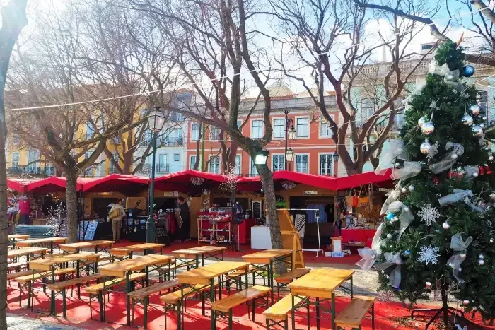 Um mercado natalino com barraquinhas decoradas e uma árvore de Natal ao fundo, em uma praça ao ar livre.
