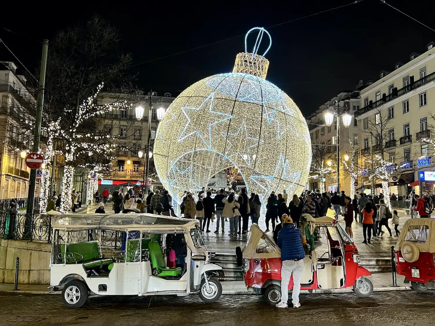 Uma grande instalação luminosa em formato de bola de Natal decorada com luzes brancas e douradas, cercada por tuk-tuks e pedestres em uma praça movimentada à noite.