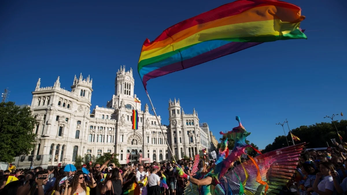 Palacio branco de Madrid ao fundo, com bandeira lgbt arco iris a frente e pessoas na parada lgbt