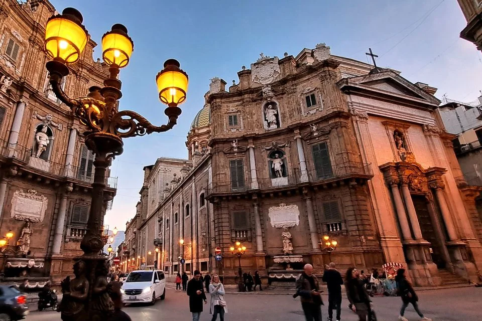 Praça com predios antigos, um poste com 3 lâmpadas a esquerda, pessoas e carros passando