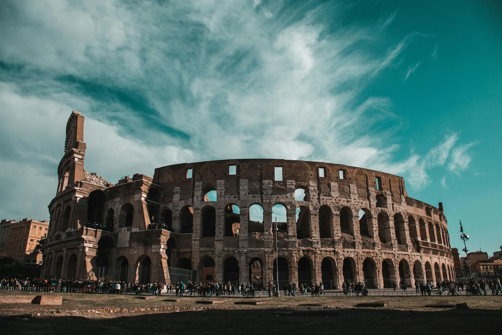 oliseu em Roma, Itália, com céu azul e algumas nuvens ao fundo. Diversos turistas se reúnem na área externa do monumento histórico.