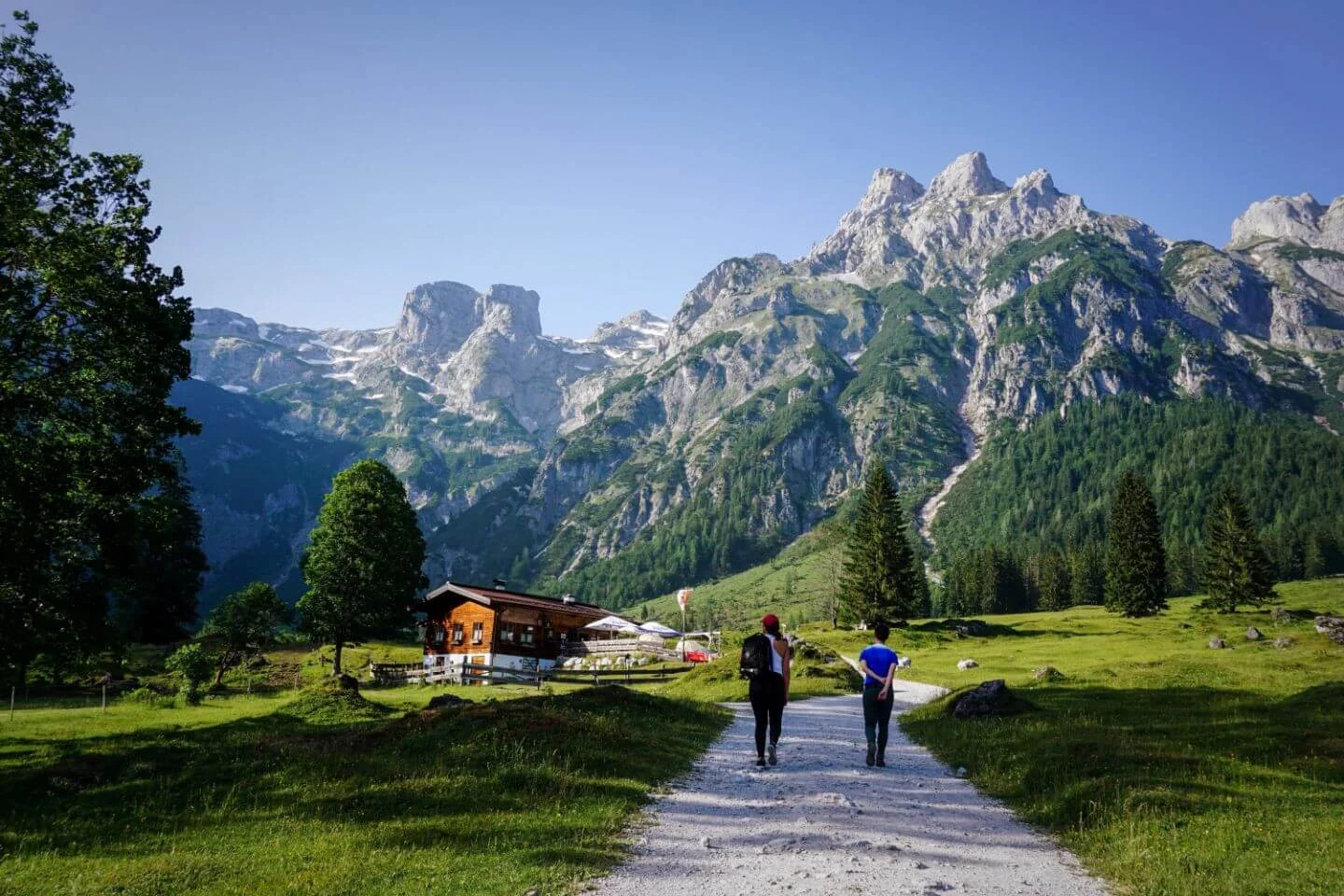 duas pessoas caminhando em um caminho de terra branco, com gramado ao lado, e montanhas ao fundo