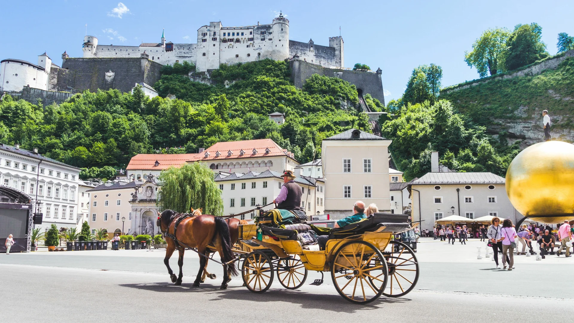 carroça amarela com dois passageiros, dois cavalos marrons, em uma praça com um castelo ao fundo