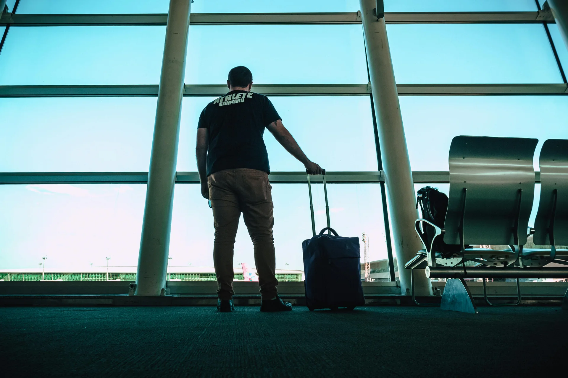 Homem olhando para uma janela de aeroporto com uma mala ao lado, esperando o voo para viagem internacional.