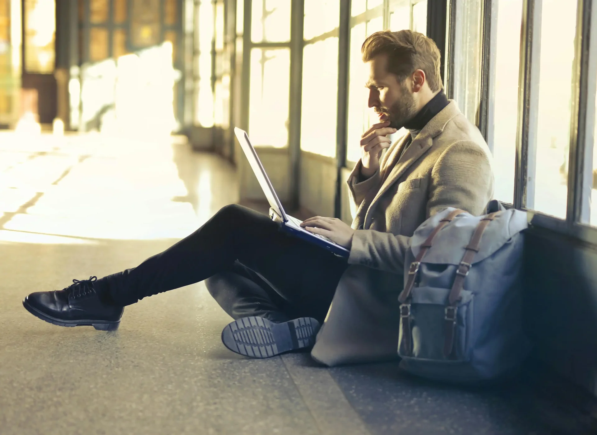 Homem sentado no chão do aeroporto, focado em seu laptop, planejando detalhes de viagem.